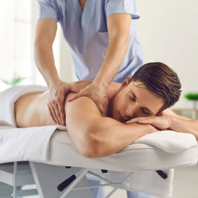 A patient lies prone on a treatment table while a massage therapist works between the shoulders